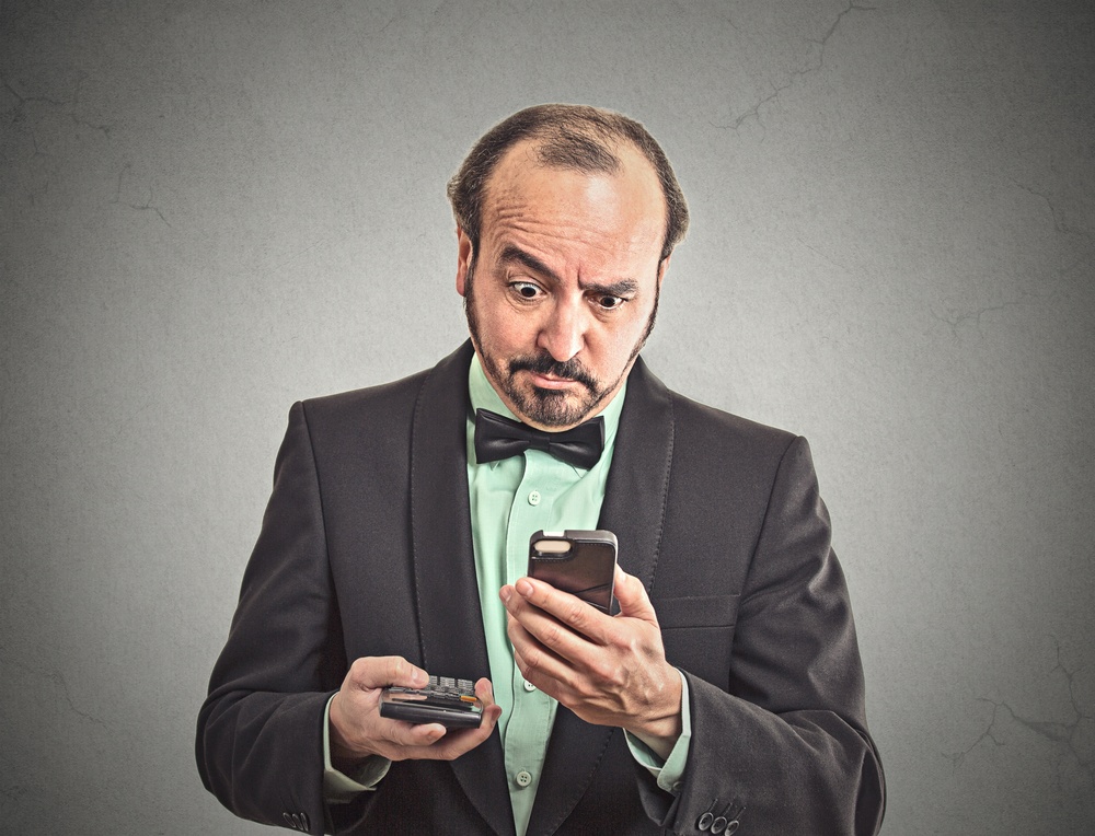 Portrait surprised businessman looking on smart phone holding calculator unexpected financial bills charges isolated grey wall background. Human face expression, emotion, body language, reaction